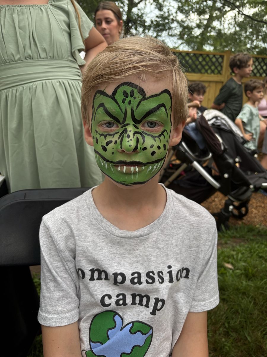 Boy wearing a full-face green dinosaur design with black outlines, painted scales and white teeth