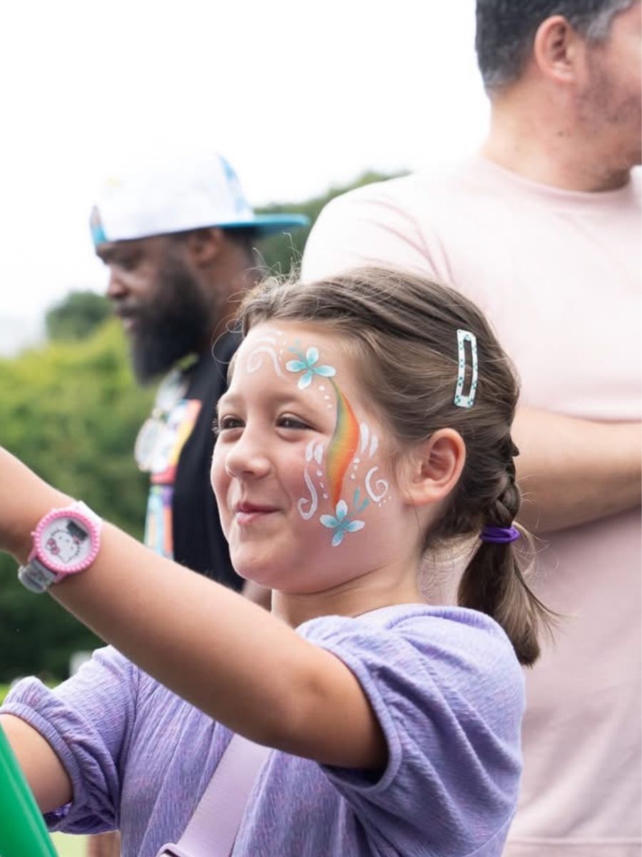 Girl smiling at an outdoor event with a pastel rainbow, flowers and swirls painted on her cheek
